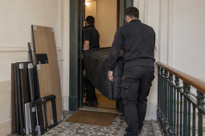 Un bureau compact sous housse zippée, sur un palier d'appartement près d'une porte étroite, déménagement de Paris vers le Burkina Faso