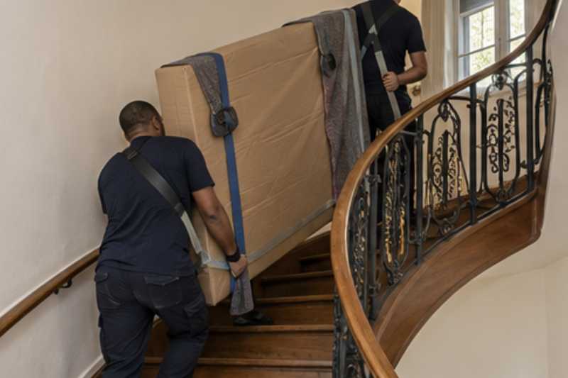 Un sommier double sous carton plat et couverture épaisse, dans un escalier tournant en bois d'immeuble ancien, déménagement de Paris vers les Fidji