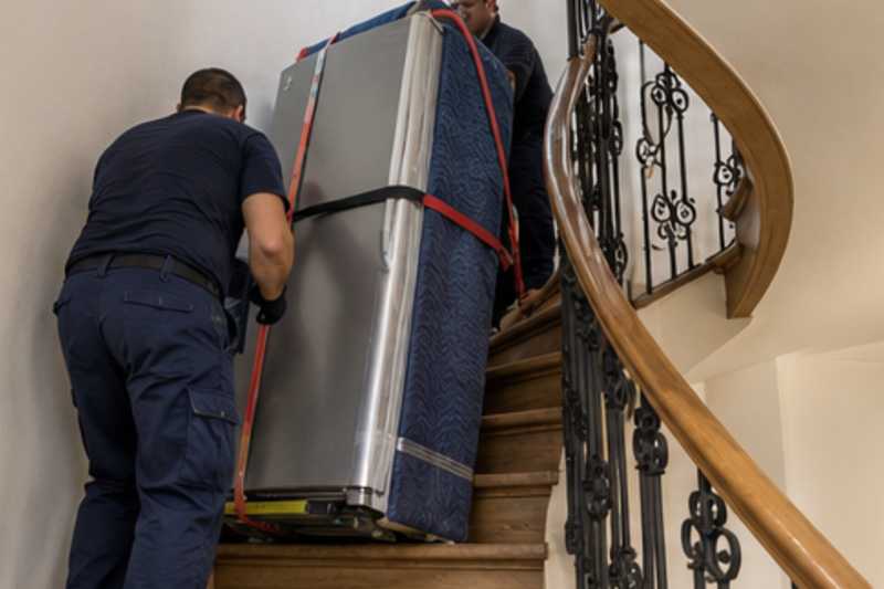 Un réfrigérateur combiné sur un diable sous couverture de déménagement, dans un escalier tournant en bois d'immeuble ancien, déménagement de Paris vers la Guinée équatoriale