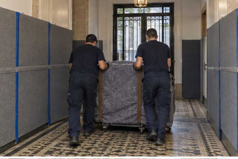 Une commode à six tiroirs sous couverture de déménagement, dans le hall d'une résidence aux murs protégés par des panneaux de déménagement, déménagement de Paris vers le Honduras