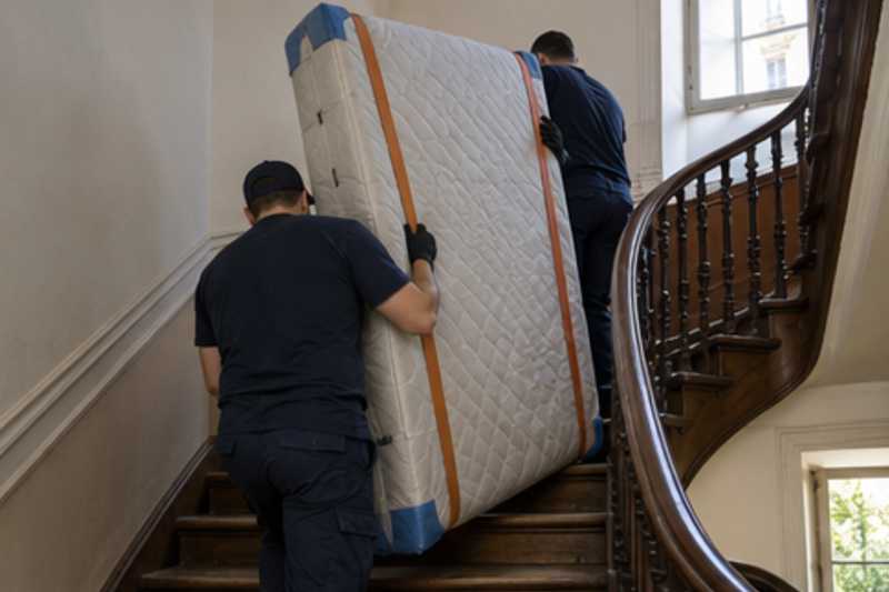 Un grand matelas debout avec protections d'angles, dans un escalier tournant en bois d'immeuble ancien, déménagement de Paris vers la Moldavie