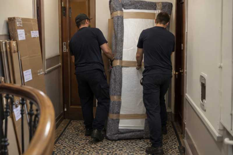 Un grand miroir encadré tenu debout sous carton plat et couverture épaisse, sur un palier d'appartement près d'une porte étroite, déménagement de Paris vers Tel-Aviv